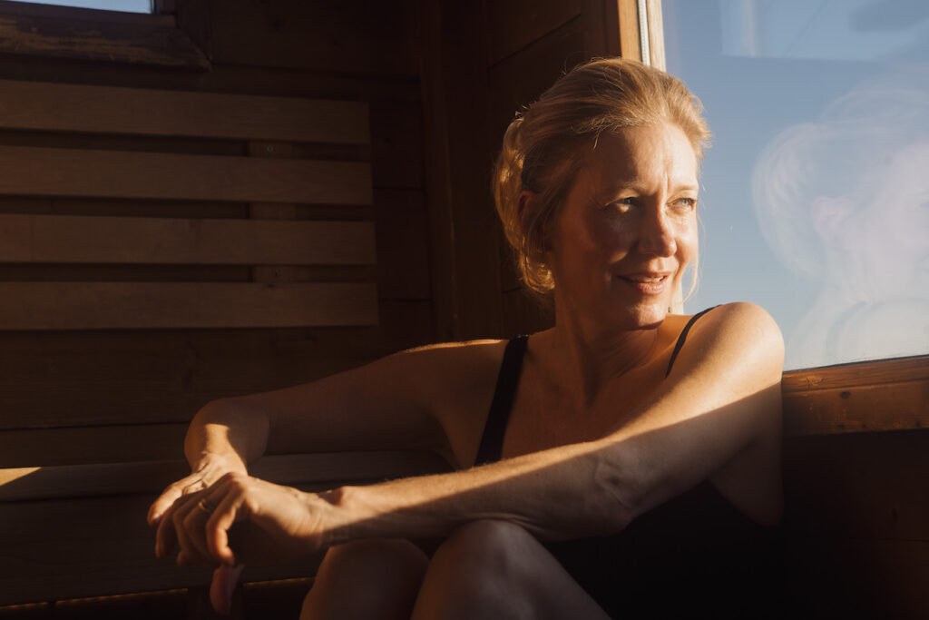 Blond kvinne ser ut av vinduet i badstuen. Smiler mot solen. Badstue